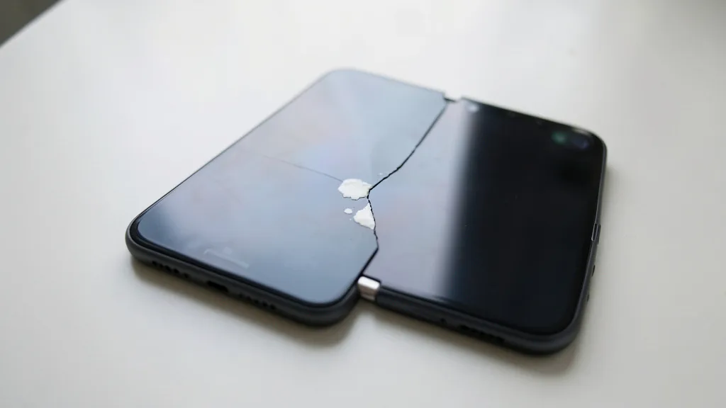 Imagem gerada por IA: Celular dobrável aberto com uma rachadura discreta e uma mancha clara na tela interna, junto à dobra.