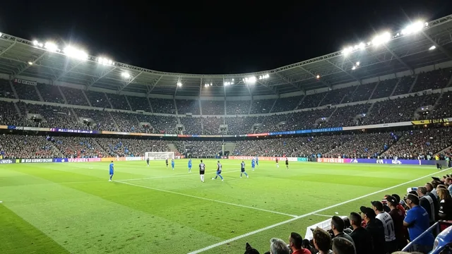 Imagem gerada por IA: Vista ampla de um estádio de futebol iluminado durante uma partida, com jogadores em campo e torcida nas arquibancadas.