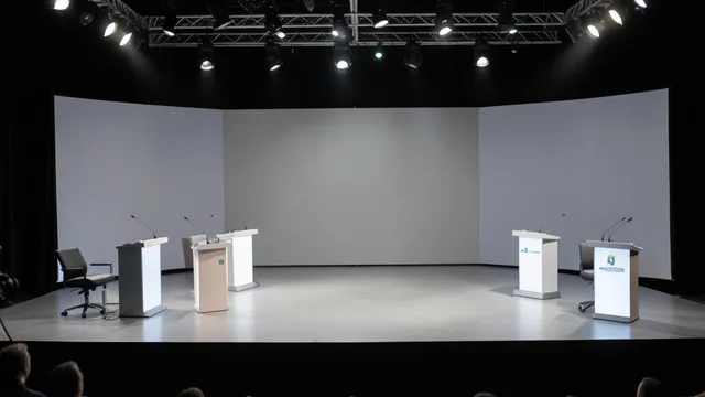 Palco de debate presidencial com púlpitos vazios e iluminação de estúdio.