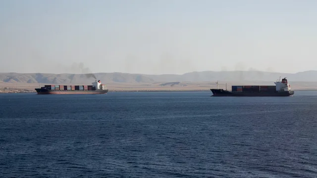 Imagem gerada por IA: Navios comerciais no mar perto de uma costa árida, com fumaça ao longe.