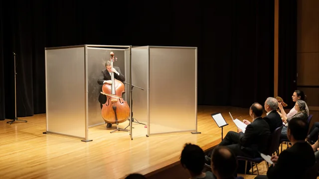Imagem gerada por IA: Contrabaixista toca atrás de um biombo durante uma audição em uma sala de concertos.