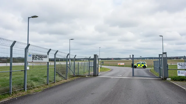 Entrada protegida de uma base aérea britânica, com cerca e viatura policial sob céu nublado.